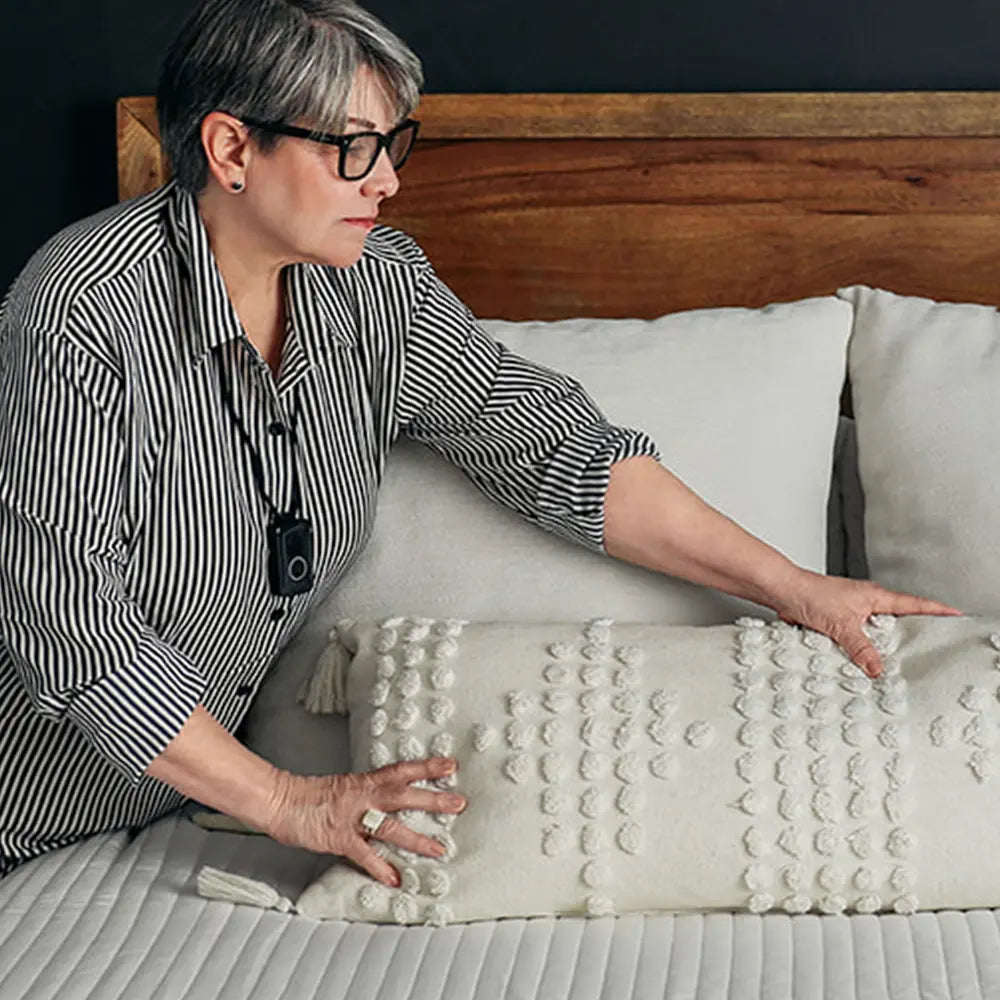 A woman arranging decorative pillows on a bed, wearing her Belle S medical alert pendant.