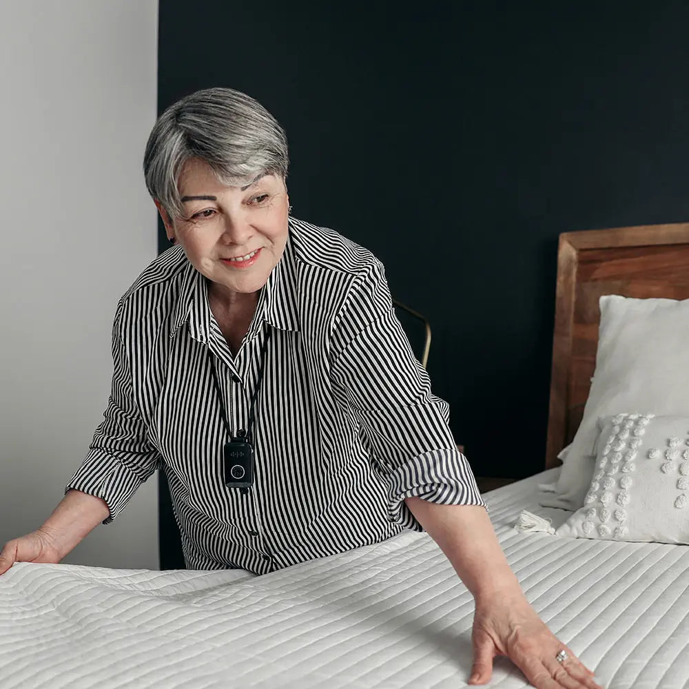 Woman in a striped shirt standing next to a bed with a medical alert device around her neck. 